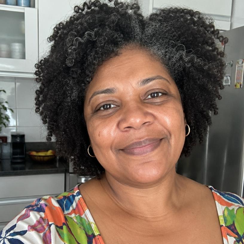 Foto de perfil de Cláudia, mulher de cabelo cacheado, sorrindo na cozinha de casa